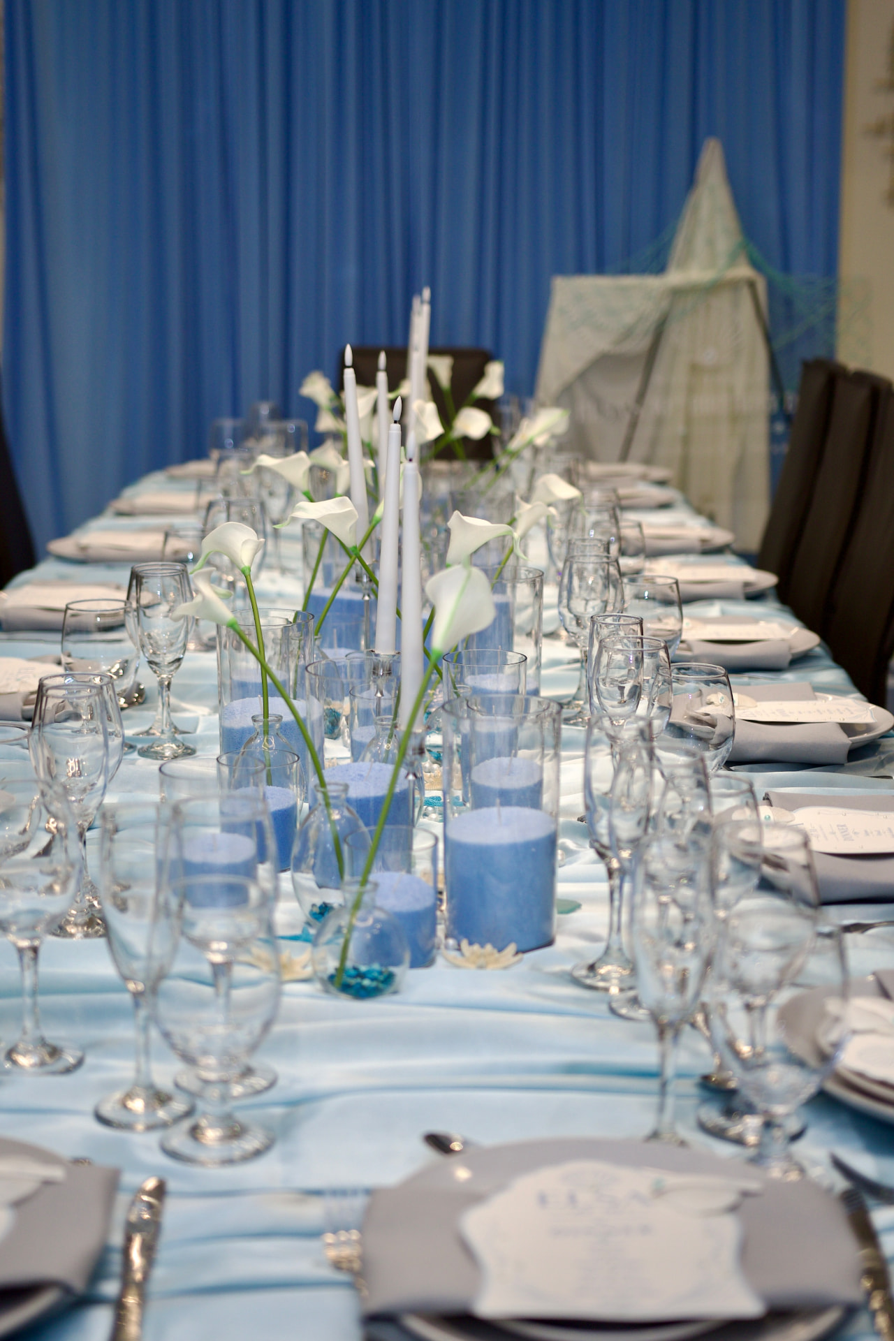 Long satin blue tablescape with calla lilies, taper candles and blue sand candles beneath draped curtains
